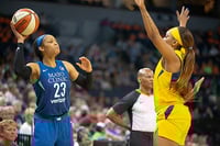 With fans out of focus in the background, a basketball player in blue, holding a ball, faces off against a player in yellow, with a referee behind her.